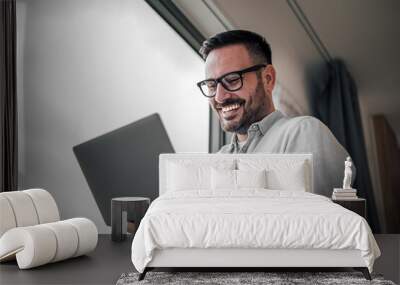 Portrait of young adult smiling cheerful businessman entrepreneur in office using laptop computer having online video call conference meeting standing next to window. Wall mural