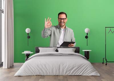 Portrait of young man showing OK sign and checking social media apps over tablet on green background Wall mural