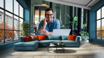 Portrait of young smiling cheerful entrepreneur in casual office making phone call while working with laptop Wall mural