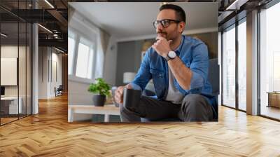 Positive male entrepreneur with hand on chin thinking business strategies while sitting comfortably on armchair. Young pensive freelancer holding coffee cup and looking away while working home office Wall mural