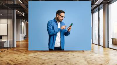 Serious handsome young adult man dressed in denim shirt surfing internet over smart phone while standing isolated against blue background Wall mural