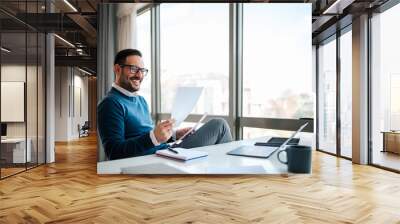 Smiling business professional reading documents while sitting at office desk Wall mural