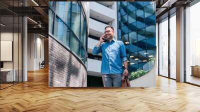 Smiling businessman talking on smart phone while walking against office building Wall mural