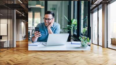 Smiling businessman using phone in home or office. Small business entrepreneur looking at his mobile phone and smiling reading or texting good news. Wall mural