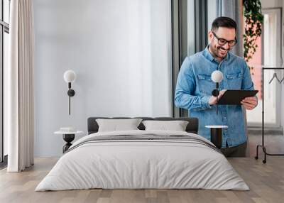 Smiling businessman wearing glasses using computer tablet, standing in modern business space. Wall mural