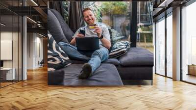 smiling caucasian man, using his credit card and laptop, at home. Wall mural