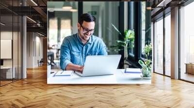 Smiling entrepreneur working on laptop while sitting at table in home office Wall mural