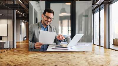 Smiling male executive reading documents while working on laptop at office desk Wall mural