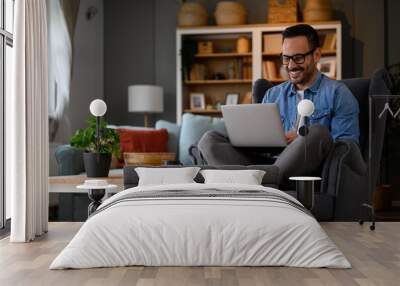 Smiling male web designers dressed in denim shirt working over laptop and sitting with crossed legs on armchair. Young businessman coding over wireless computer while working from home office Wall mural