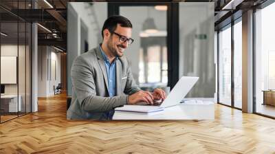 Smiling young entrepreneur typing on laptop while working at office desk Wall mural