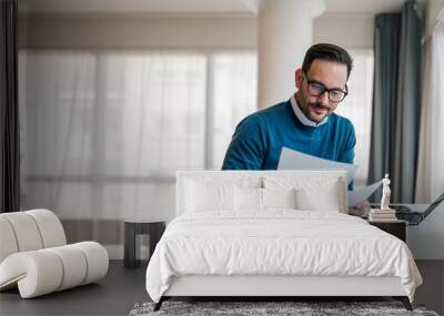 Smiling young executive analyzing business documents. Male professional is working on laptop. He is wearing formals while sitting at office desk. Wall mural