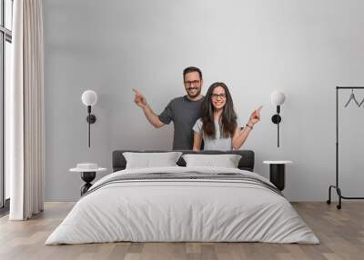 Smiling young man and woman pointing at copy space for advertisement on background. Happy couple wearing eyeglasses and casuals looking at camera and showing empty space for marketing Wall mural