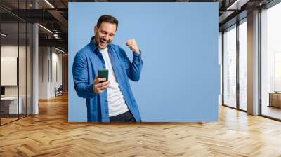 Successful male entrepreneur pumping fist after reading good news over cellphone on blue background Wall mural