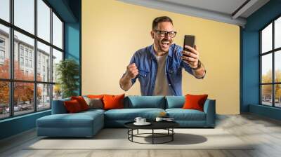 Successful male freelancer reading good news over mobile phone and screaming cheerfully against beige background. Young man dressed in denim shirt pumping fist while messaging on cellphone Wall mural