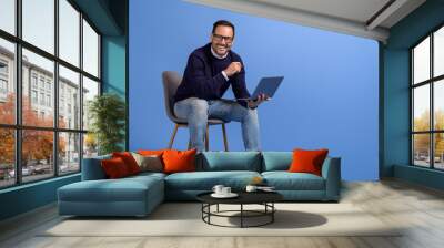 Successful male manager with laptop smiling at camera while sitting on chair over blue background Wall mural