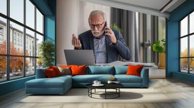 Worried senior businessman talking on mobile phone while using laptop at desk Wall mural