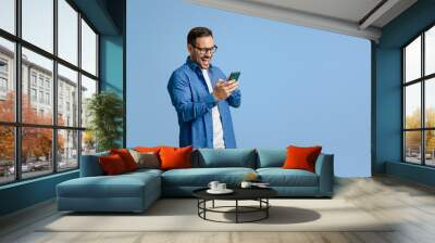 Young businessman laughing ecstatically while reading messages over mobile phone on blue background Wall mural