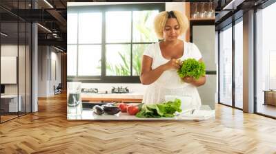 Young African American curvy woman standing in the kitchen preparing a salad of green lettuce leaves, different vegetables on the table. Diet and healthy lifestyle. Body positive. Wall mural