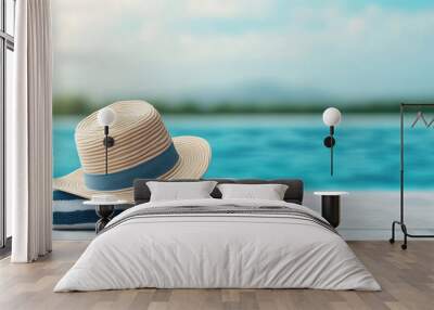 A stylish straw hat rests on a blue striped towel by a serene pool, evoking relaxation and summer vibes. Wall mural