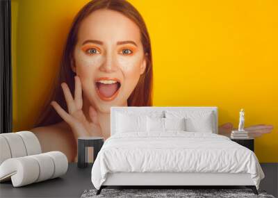 Beautiful girl with shiny brown straight long hair. Woman with orange makeup with freckles. Girl near empty copy space shows product . Girl surprised, on bright orange background Wall mural