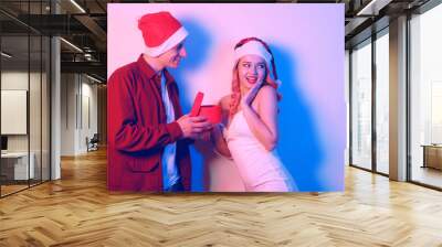 Couple in santa hats, new year, christmas. Lovers in neon light. Guy gives a gift in a red box to a happy girl, surprised woman in shock Wall mural