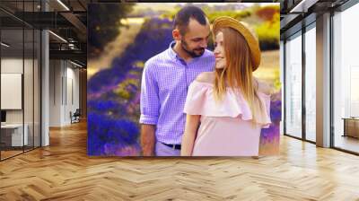 Cute young happy couple in love in a field of lavender flowers. Enjoy a moment of happiness and love in a lavender field. Boy smile to his girl. Blond girl in hat and pink dress. Romantic couple. Wall mural