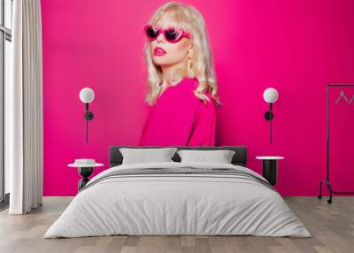 The blonde poses on a pink background in stylish pink glasses and a dress. Girl model posing, bright photo fashion and style, gold jewelry 80s and 90s Wall mural