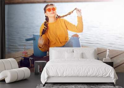 The girl at hot sunny day have fun. Summer vibes. Girl with fashion yellow sunglasses and dressed in yellow. Beautiful girl sitting on the pier near the river. Beauty model with lemonade and camera. Wall mural