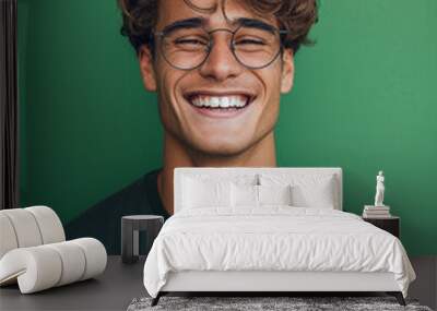 Confident young man with stylish glasses, smiling in casual attire on green background Wall mural