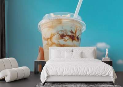 Hand holding iced coffee against blue sky backdrop. Refreshing summer drink with creamy swirls. Perfect for hot days or a caffeine boost. Wall mural