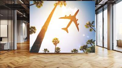 Low angle view of a plane flying in the sky surrounded by palm trees Wall mural