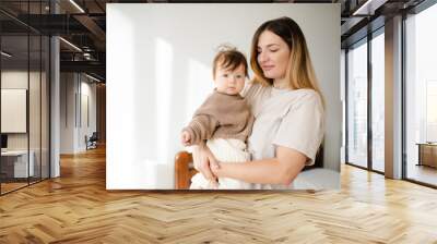 Cute infant baby girl under 1 year old on mother hands wear casual knit sweater and pants in bedroom. Look at camera. Motherhood. Wall mural