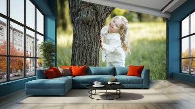 Smiling cute child girl 2-3 year old with blonde hair hide behind tree in meadow with green grass outdoor Summer season. Childhood. Wall mural