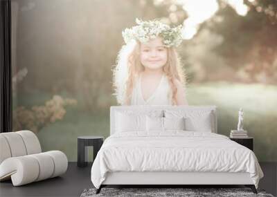 Smiling kid girl 4-5 year old wearing stylish white dress and flower wreath outdoors. Looking at camera. Sunny day.  Wall mural