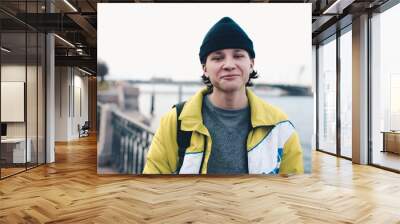 Stylish handsome guy teenager boy 18-19 year old wear knit hat and trendy winter jacket posing over river bank at city view outdoor. Smiling happy student young man with backpack. Wall mural