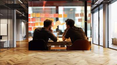 In the image, two men are sitting at a table with a laptop. They are looking at a wall covered in sticky notes, which contain notes and reminders Wall mural