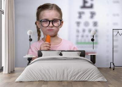 Lovely girl in glasses chewing carrot, vitamin A for good vision, healthcare Wall mural