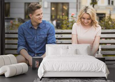 Shy blonde girl smiling, attractive guy flirting with beautiful woman on bench Wall mural