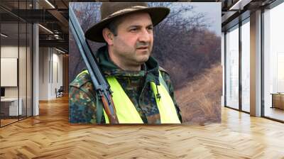 A man with a gun in his hands and an green vest on a pheasant hunt in a wooded area in cloudy weather. Hunter with dogs in search of game. Wall mural