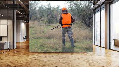 A man with a gun in his hands and an orange vest on a pheasant hunt in a wooded area in cloudy weather. Hunter with dogs in search of game. Wall mural