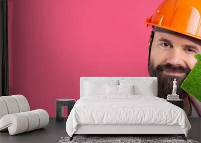 Portrait of young construction engineer wear orange hard hat, in a pink jacket standing on red studio background. A man holds a green eco house. Wall mural