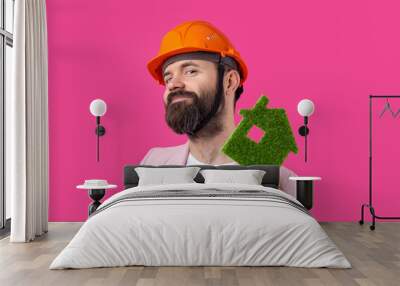 Portrait of young construction engineer wear orange hard hat, in a pink jacket standing on red studio background. A man holds a green eco house. Wall mural