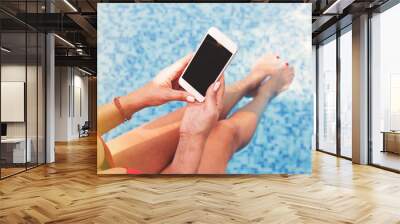 Charming woman in red swimming suit sitting on side of pool using smartphone in sunlight Wall mural