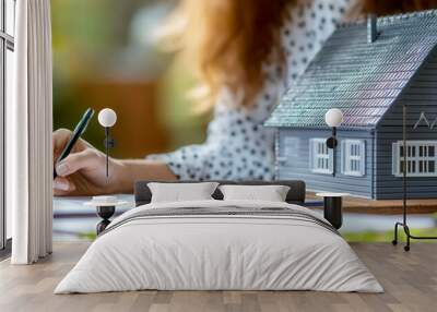 A person writes notes on a clipboard next to a miniature house model, symbolizing home ownership and real estate investment. Wall mural