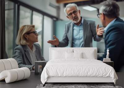 A senior professional stands explaining a point to seated colleagues in an office meeting room, gesturing with his hand, while the team listens attentively. Wall mural