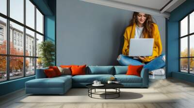 A young woman in a yellow sweater and jeans sits on the floor with her legs crossed, engrossed in her laptop. Wall mural