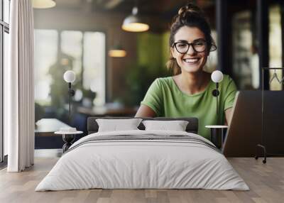 Cheerful professional woman wearing glasses and a green blouse is sitting at a desk with a laptop and holding coffee cup Wall mural