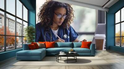 female medical professional wearing scrubs and glasses, focused on writing notes in a book Wall mural