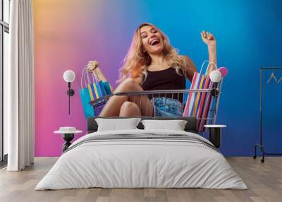 joyful woman with a raised fist, sitting in a shopping cart, holding colorful shopping bags Wall mural