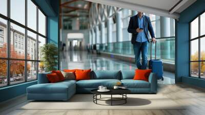 Man is walking through an airport terminal with a blue suitcase, looking at his phone. Wall mural